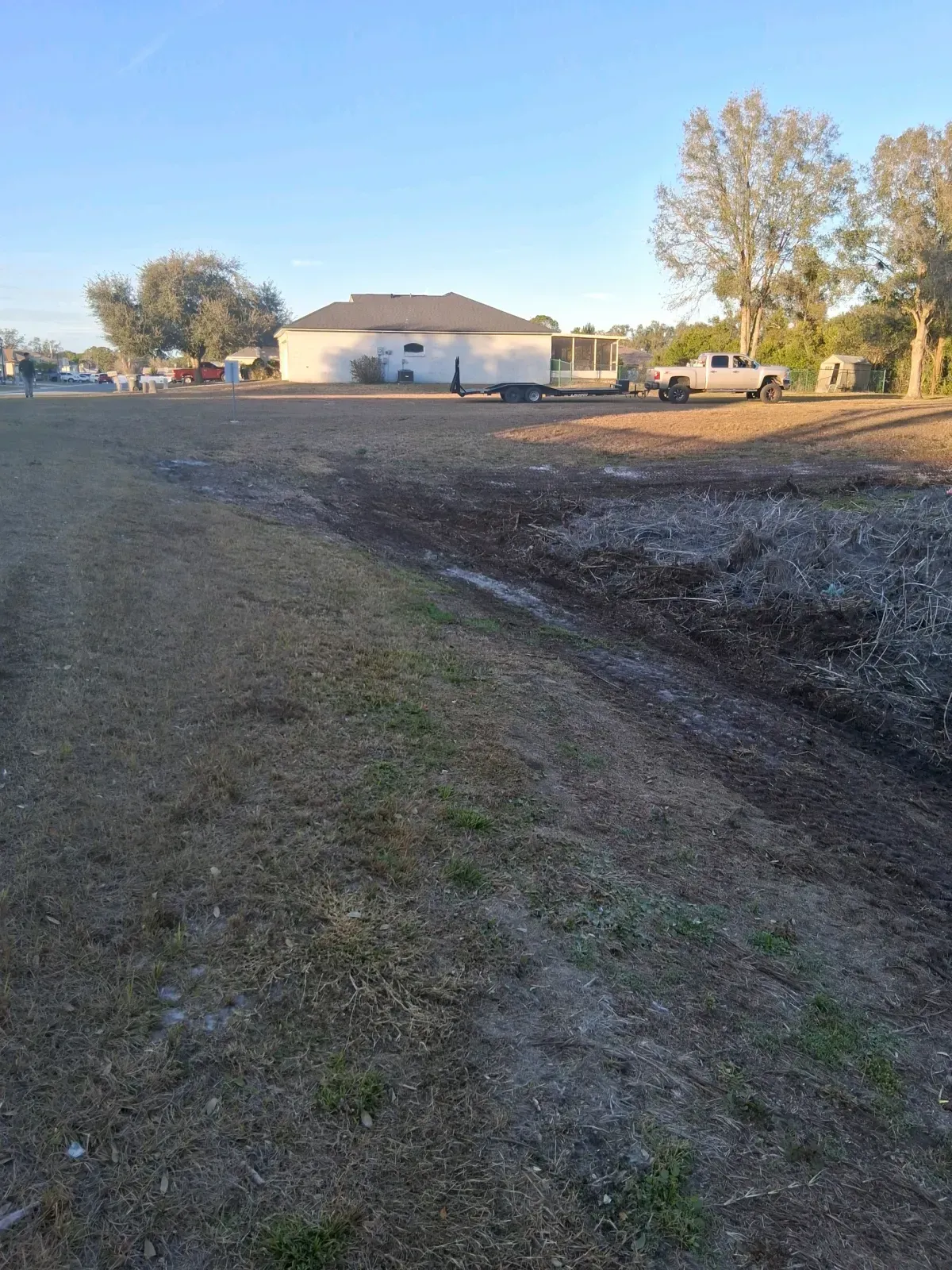 A wide view of a residential backyard with a white house, a pickup truck, and a large mound of dirt under a clear sky.