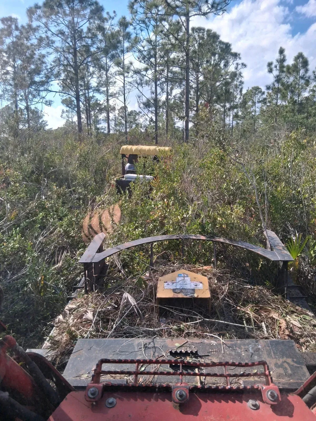 View from the cab of a tractor clearing dense brush in a forest with a utility vehicle visible ahead.