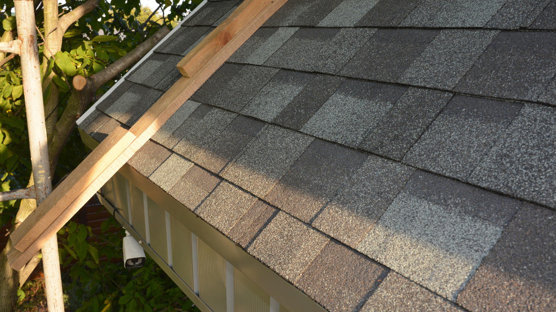 A section of a roof being worked on, featuring gray asphalt shingles and a wooden board resting against the eaves.