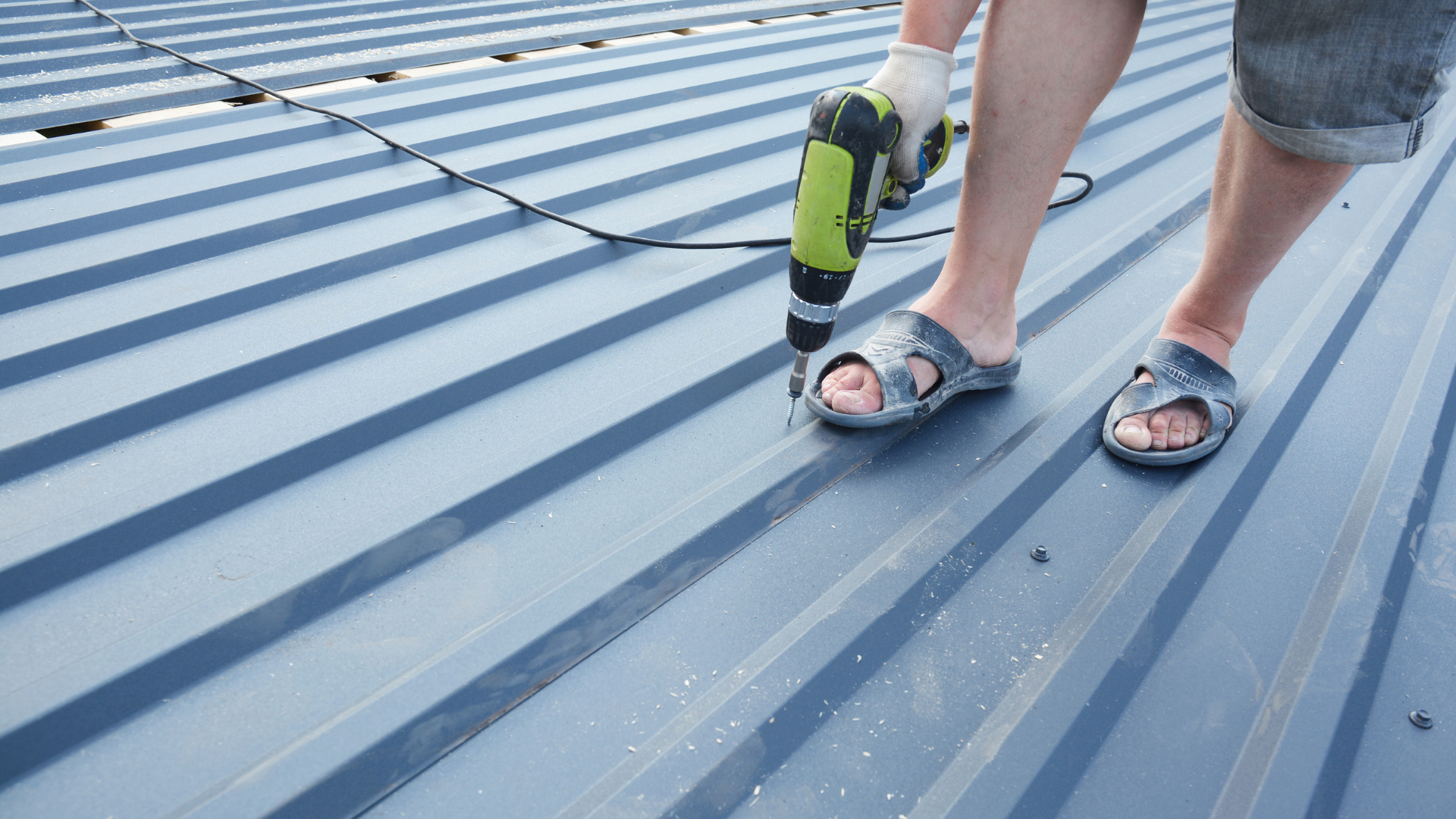 A person in sandals uses a power drill on a blue, corrugated metal roof.