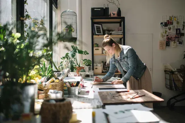 Woman in denim jacket, drawing at sunlit desk, surrounded by plants and art supplies.