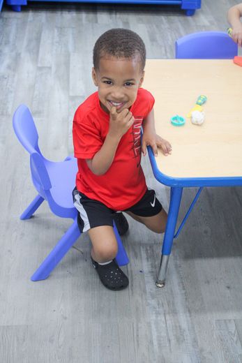 A young boy in a red shirt is sitting at a table with a blue chair - Dyer, IN - Bright Beginner's Academy
