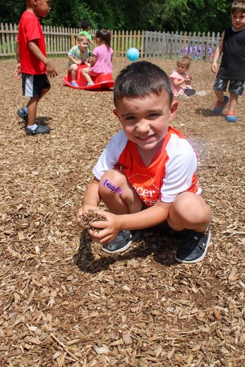 A young boy is kneeling on the ground holding a piece of wood chips - Dyer, IN - Bright Beginner's Academy
