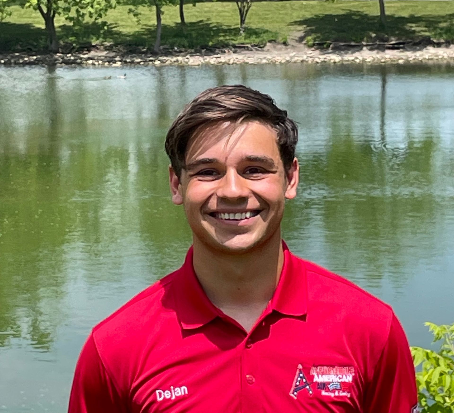 A young man in a red shirt is smiling in front of a lake.