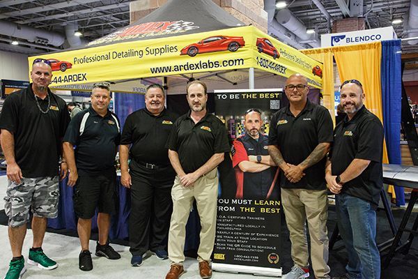 Group of seven people posing in front of a yellow trade show booth for structural design services