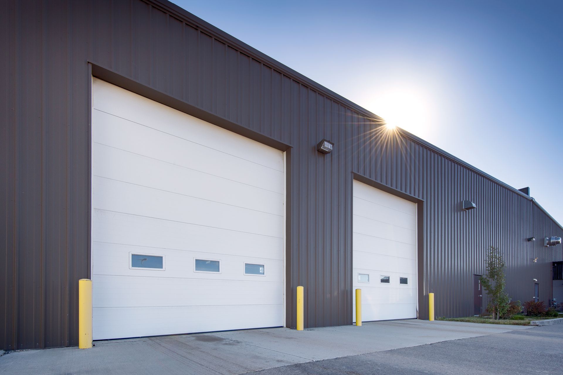A brown metal warehouse building with two large white roll-up garage doors under a bright, sunny sky.