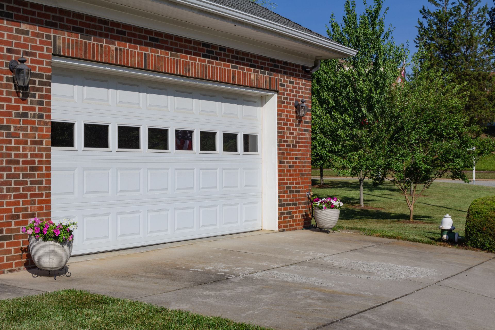 A white garage door with rectangular windows, set in a brick wall with planters on a concrete driveway.