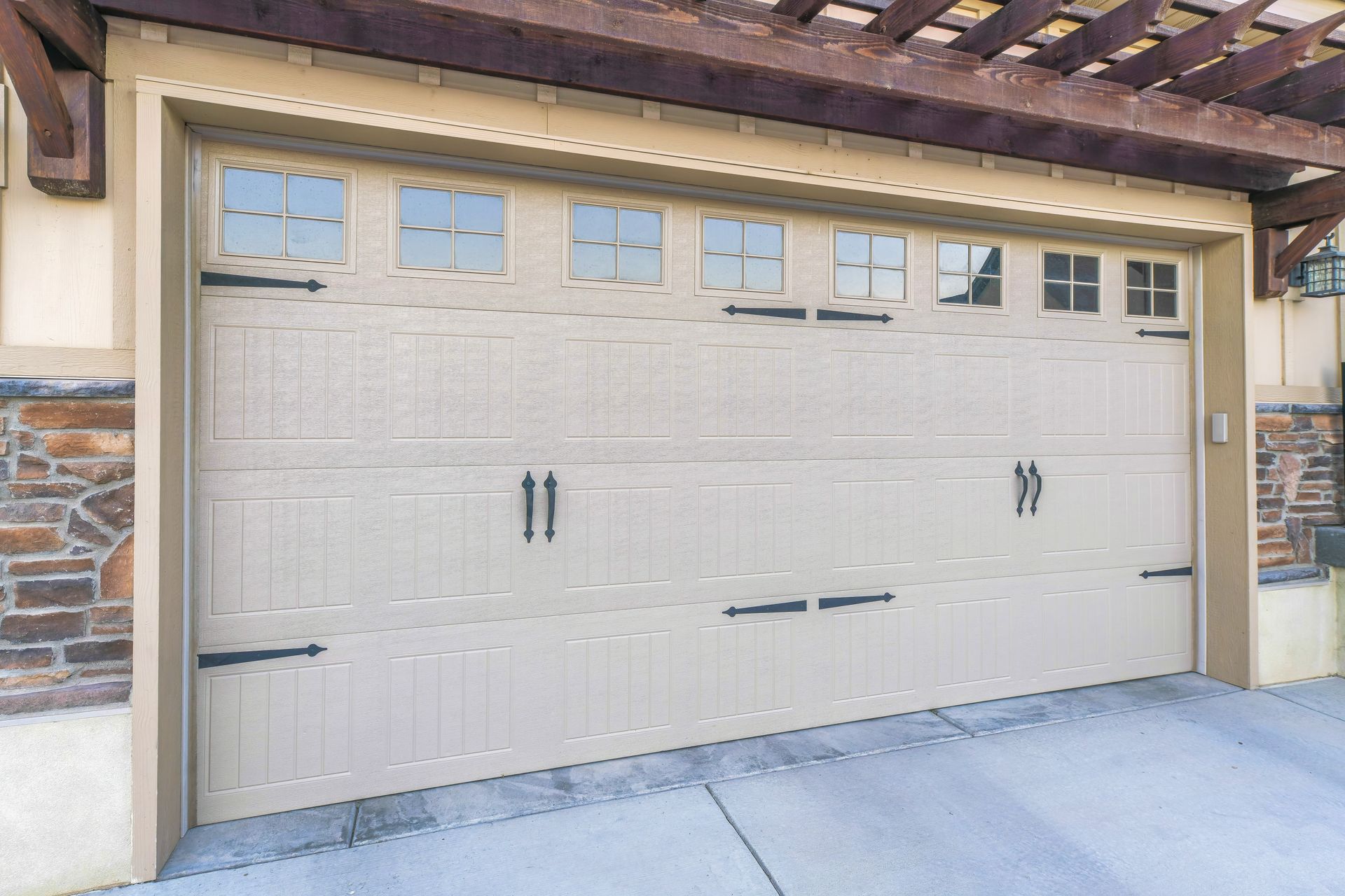 A tan garage door with decorative black hinges, handles, and small window panes in a stone-accented home exterior.