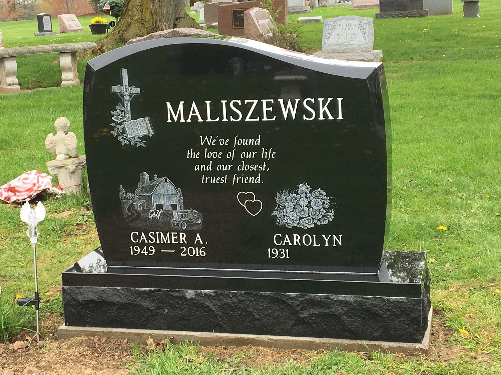 Black granite headstone with engraved names and dates, a cross, house, and trees in a cemetery.
