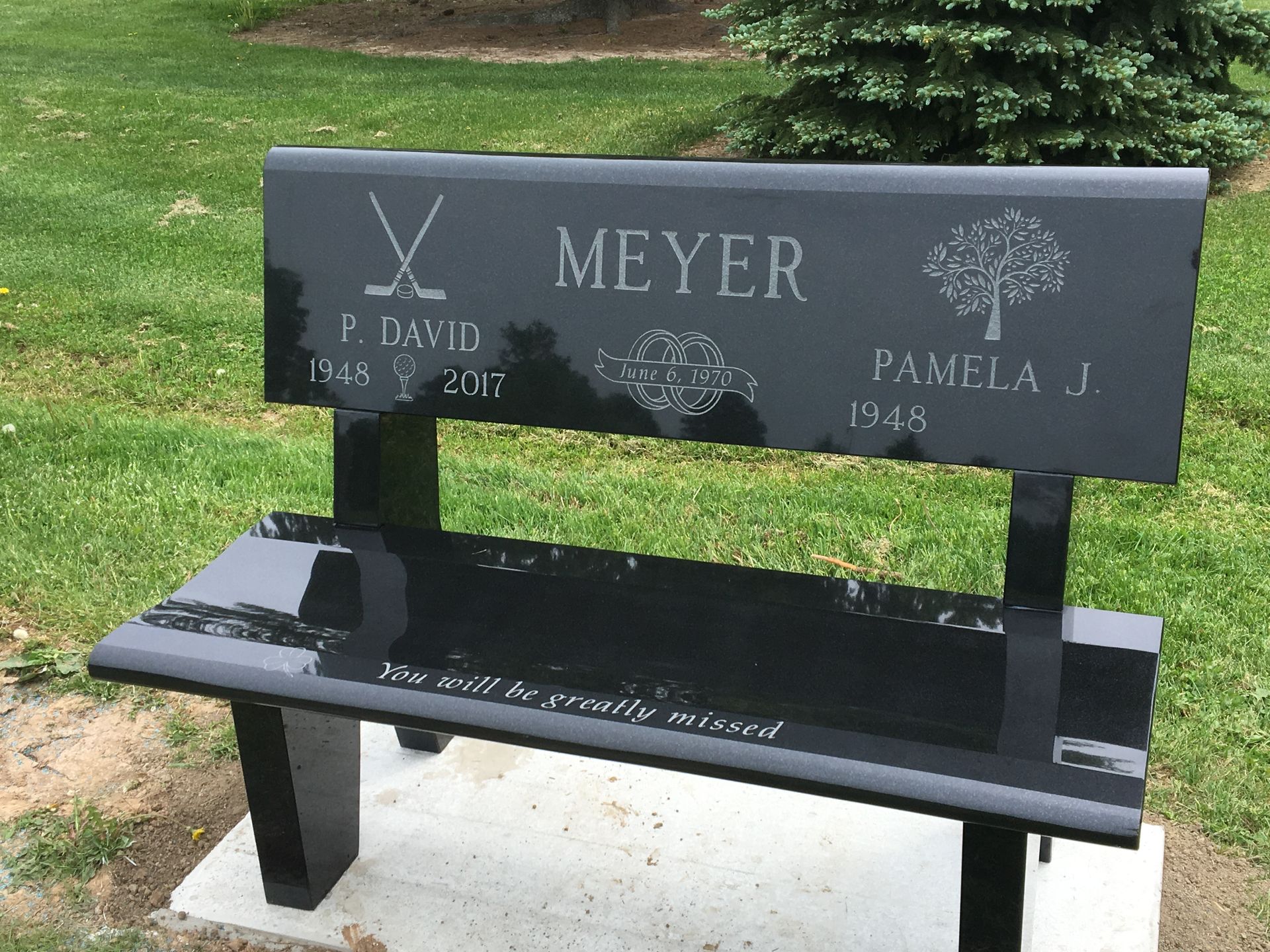Black granite memorial bench with engraved names, dates, and sports symbol in a grassy setting.