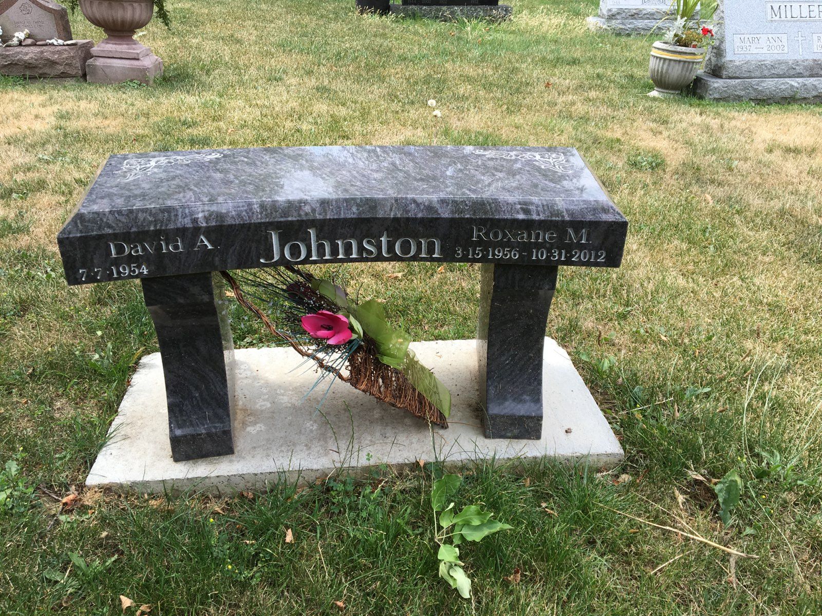 A stone bench gravestone in a cemetery; a floral arrangement rests beneath it.