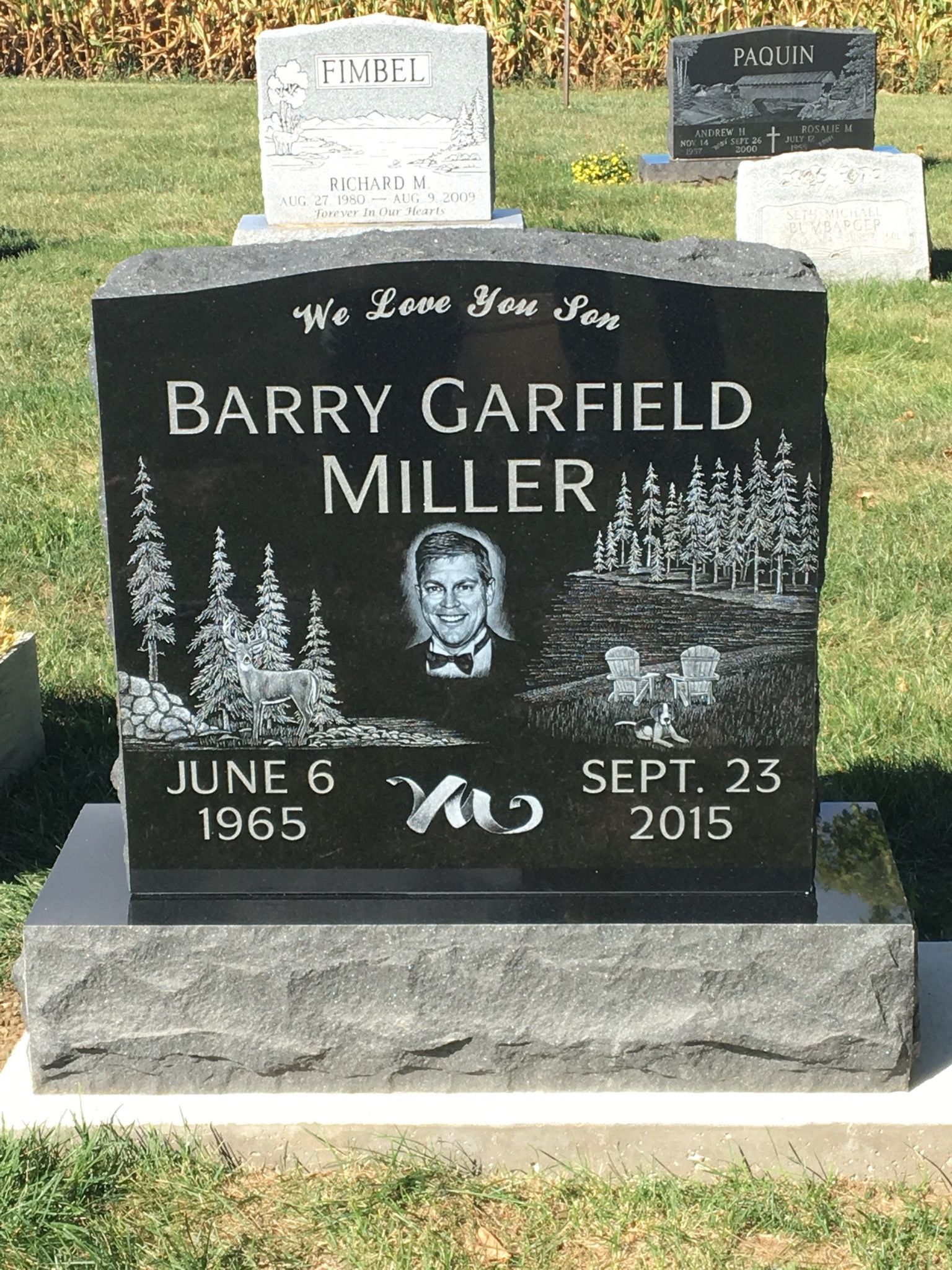 Black headstone with Barry Garfield Miller's name, dates, and image etched on it, in a grassy cemetery.
