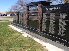 A black granite World War II memorial with names, set on a grassy median near a road.