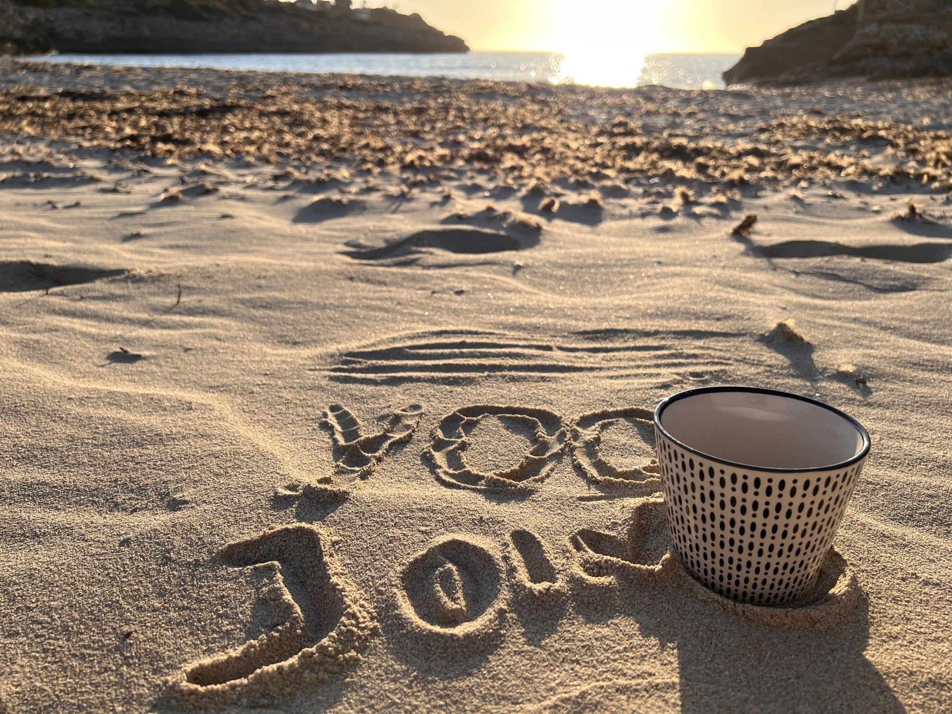 Kaffeetasse am Sandstrand bei Sonnenaufgang. Im Sand geschrieben: 