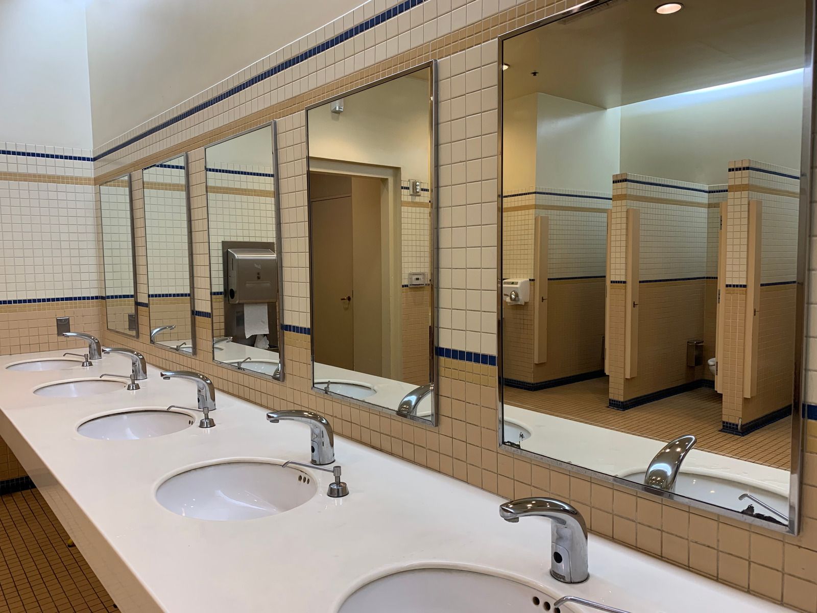 Public restroom with white sinks, mirrors, and beige tiled walls and stalls.