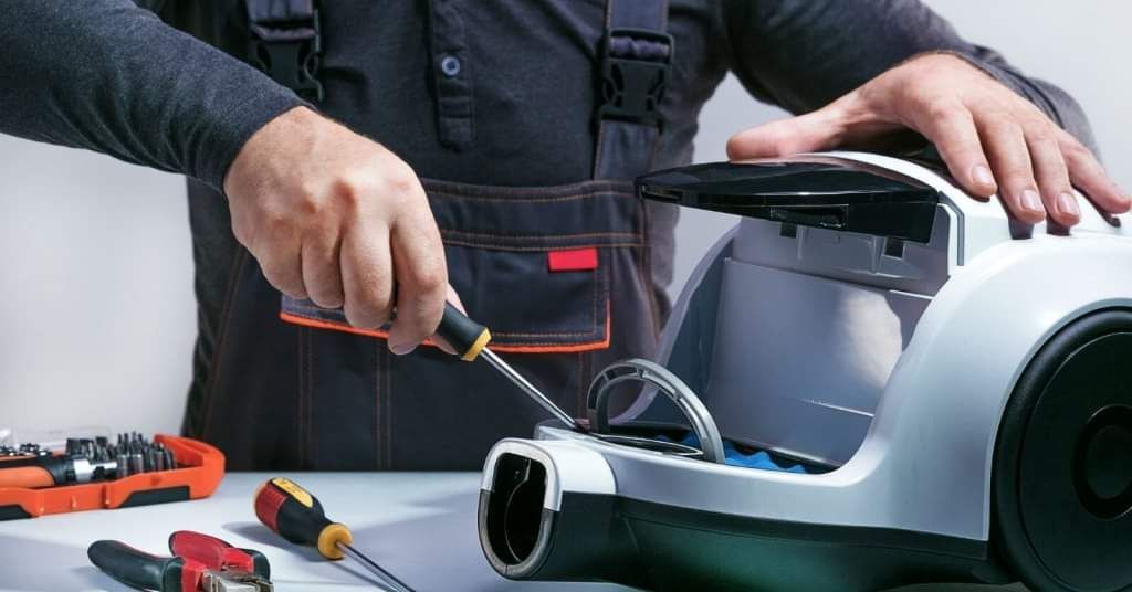 Person repairing a white vacuum cleaner with a screwdriver; tools on a table.