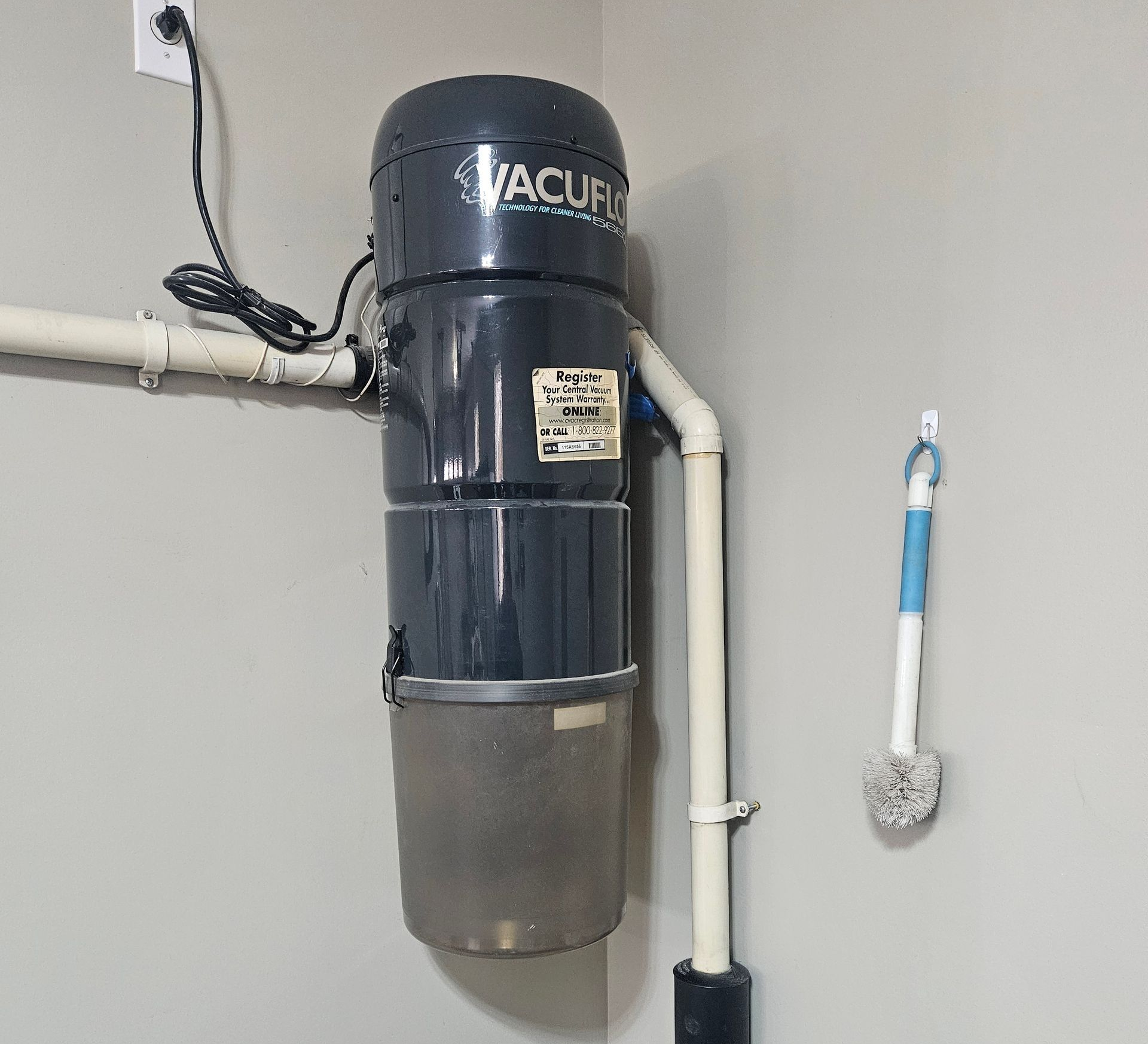 Central vacuum cleaner mounted on a light gray wall in a corner. The unit is black and cylindrical.
