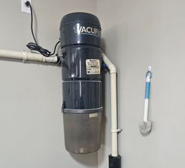 Central vacuum cleaner mounted on a light gray wall in a corner. The unit is black and cylindrical.