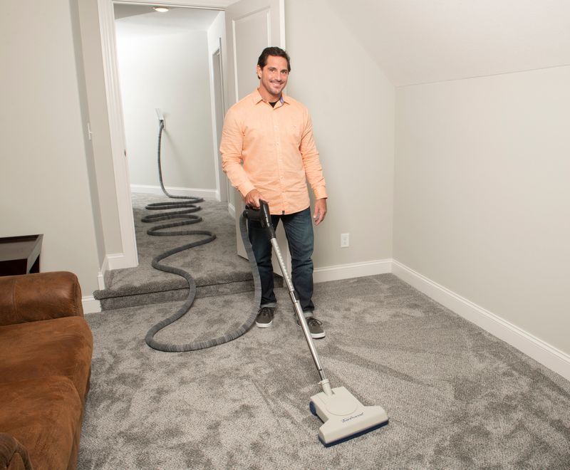 Man vacuums carpeted room with central vacuum hose trailing to hallway.