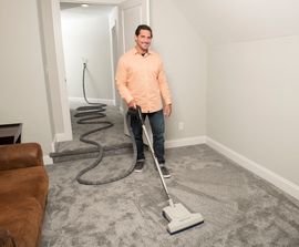 Man vacuums carpeted room with central vacuum hose trailing to hallway.