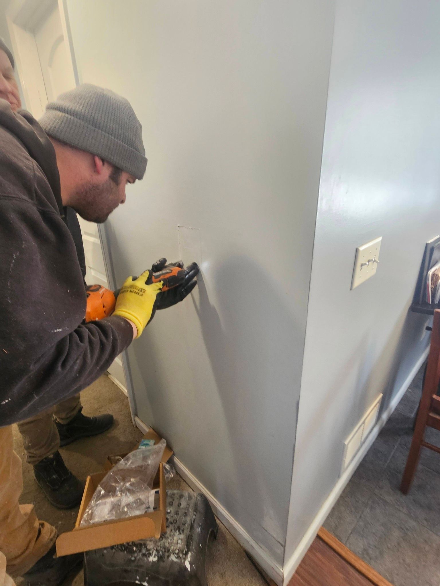 Person in beanie and gloves drilling a hole in a gray wall