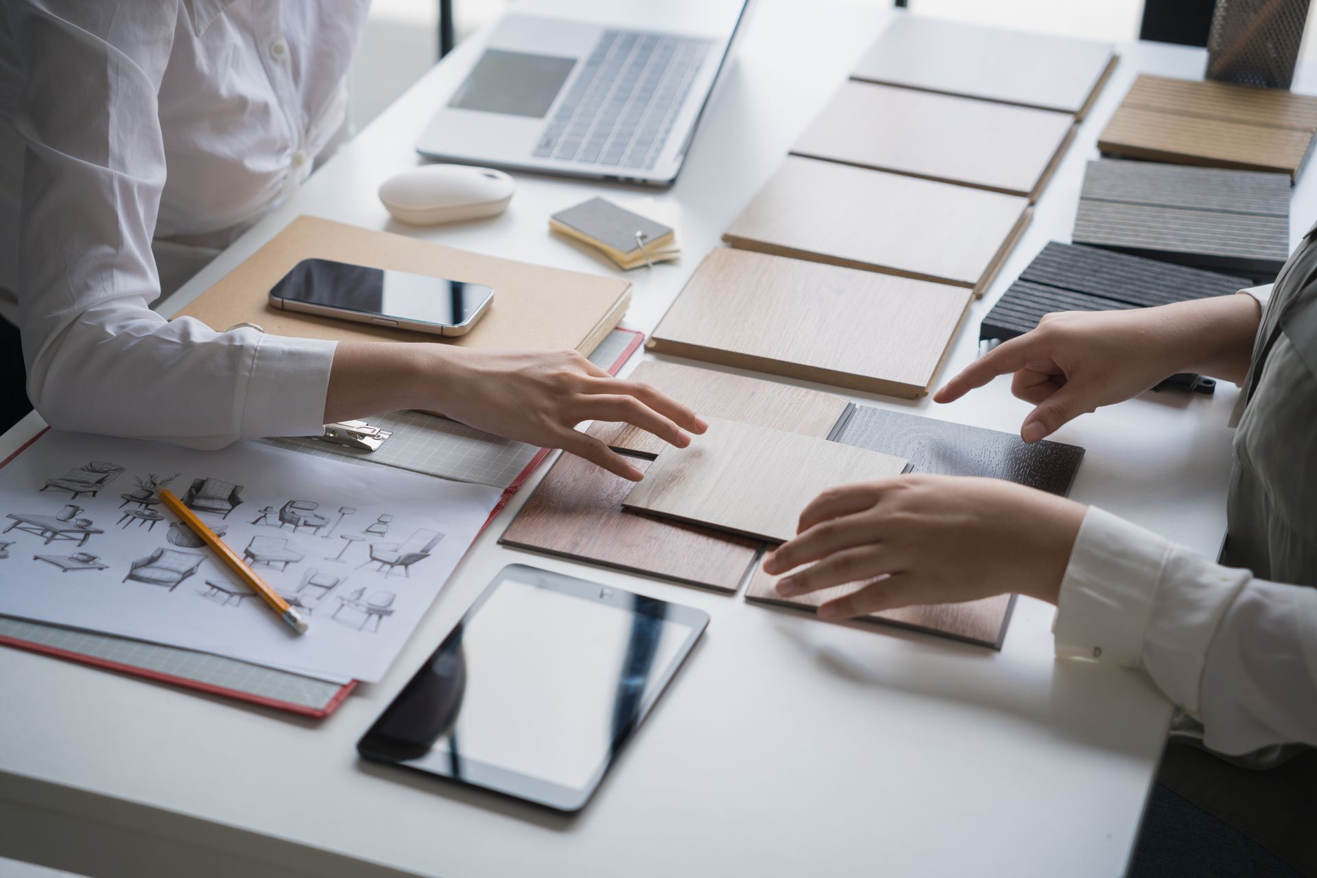 Two people reviewing material samples at a table, discussing design options with sketches, a laptop, and a tablet.