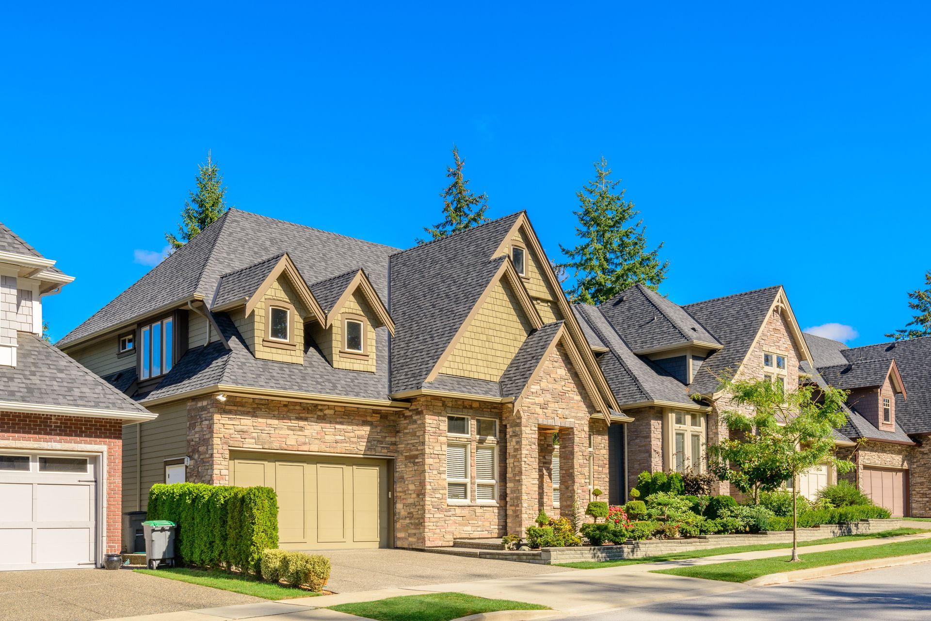 Row of brick houses with gray roofs, tan garage doors, and lush landscaping against a clear blue sky.