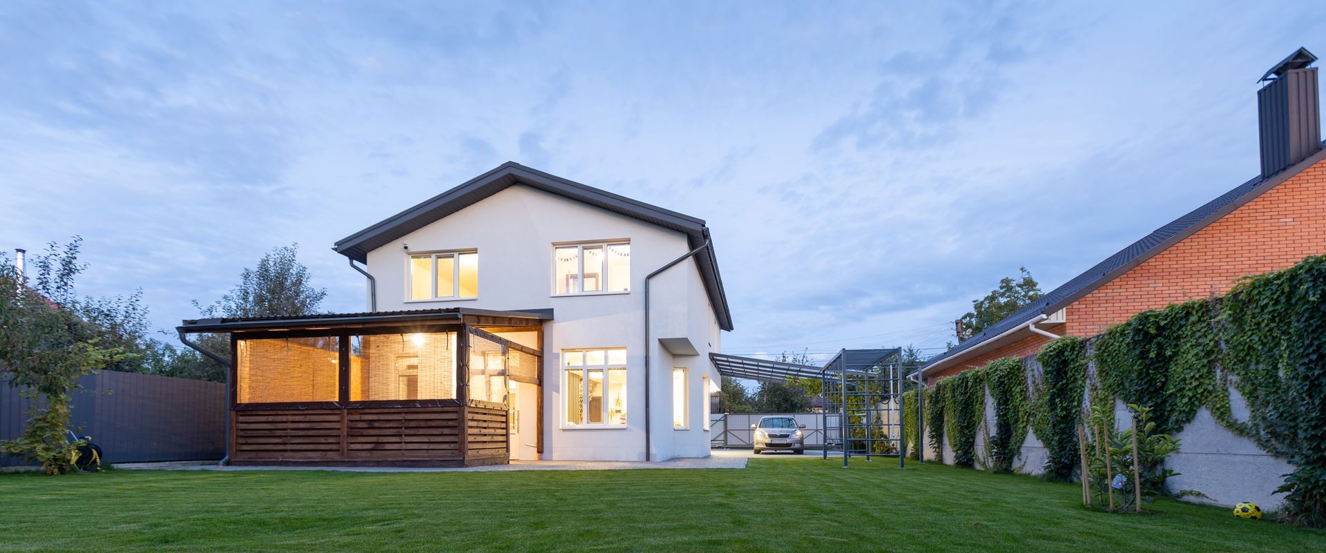 Two-story white house with lit windows and a wooden porch, green lawn, and ivy-covered brick wall.
