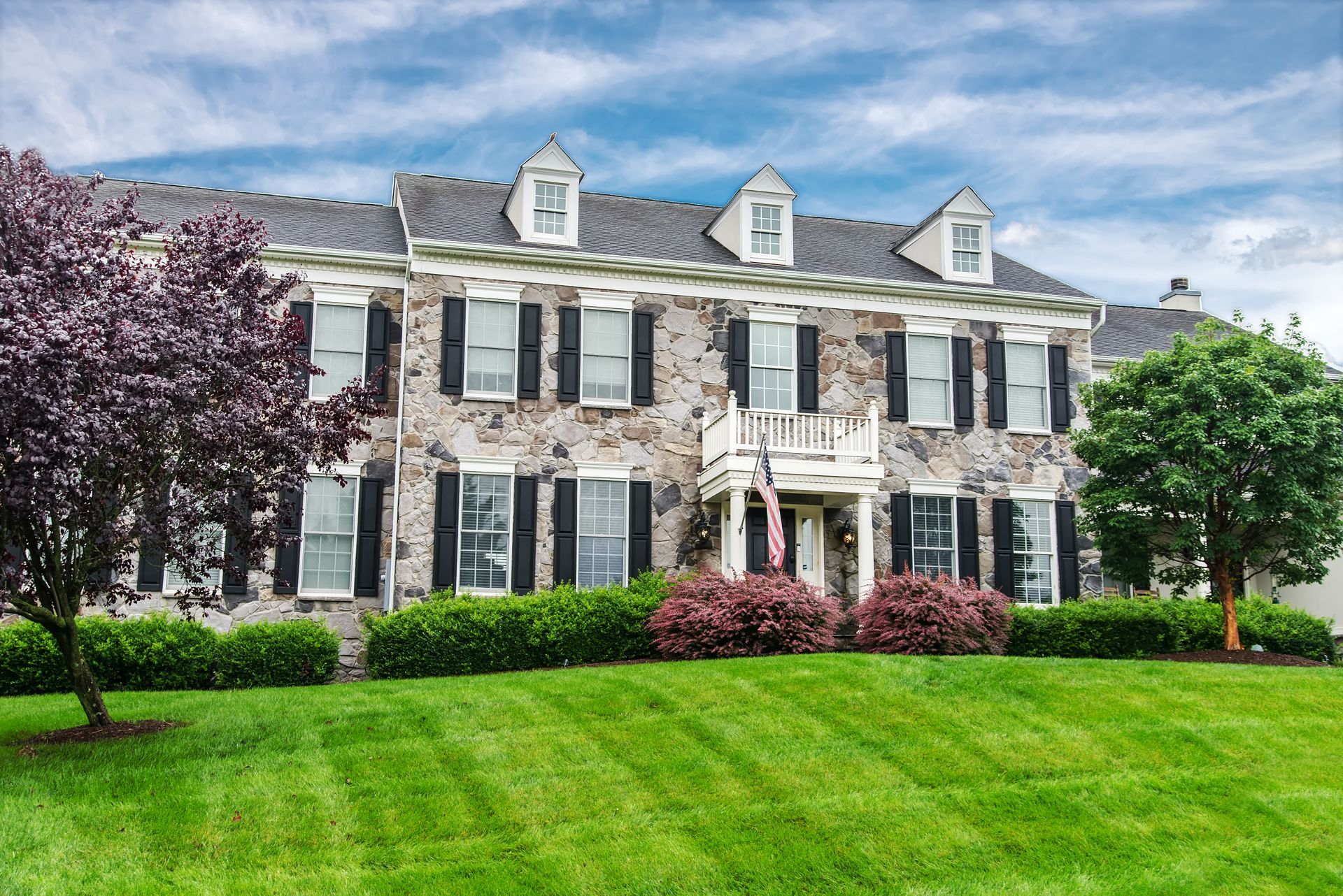 Stone house with black shutters, dormers, and a small balcony, set on a manicured lawn with trees and bushes.