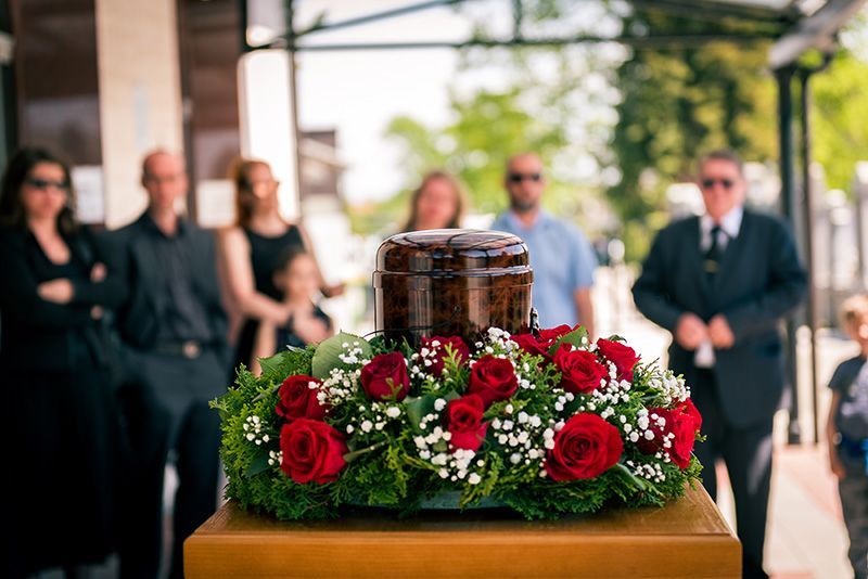 A dark urn rests atop a wooden podium, surrounded by a floral arrangement of red roses, with mourners blurred in the background.