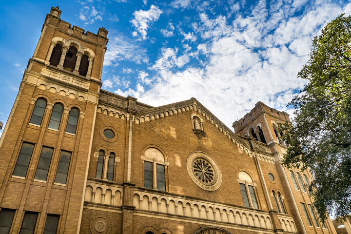 Low-angle view of a brown brick church with two towers under a blue sky with white clouds.