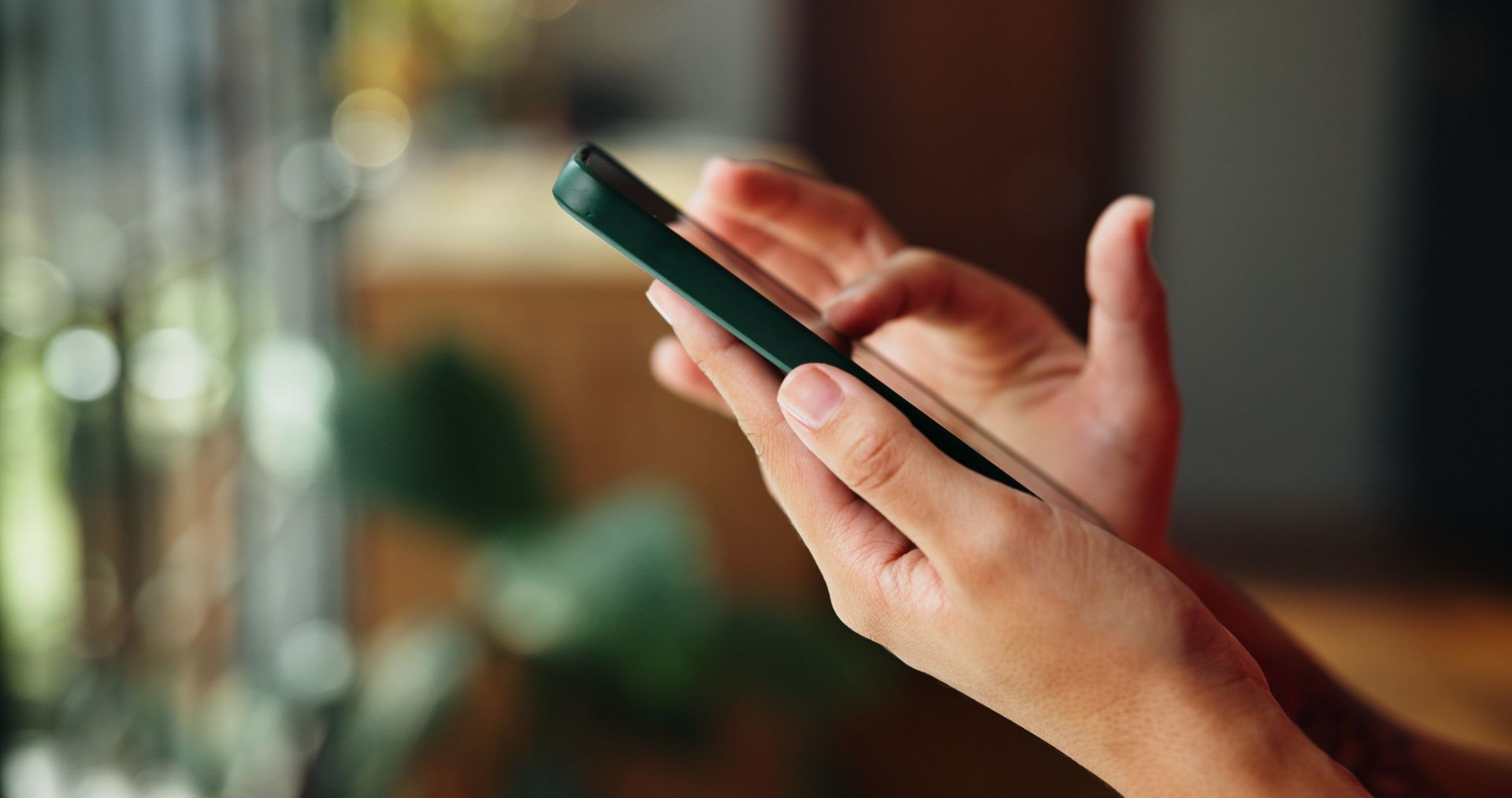 Close-up of hands holding a dark-cased smartphone in a softly blurred, warm indoor setting.