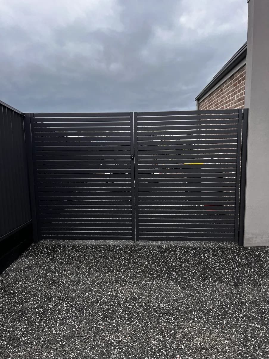 A black gate is sitting on top of a gravel driveway next to a brick building — Mid Coast Glass Fencing In Forster, NSW