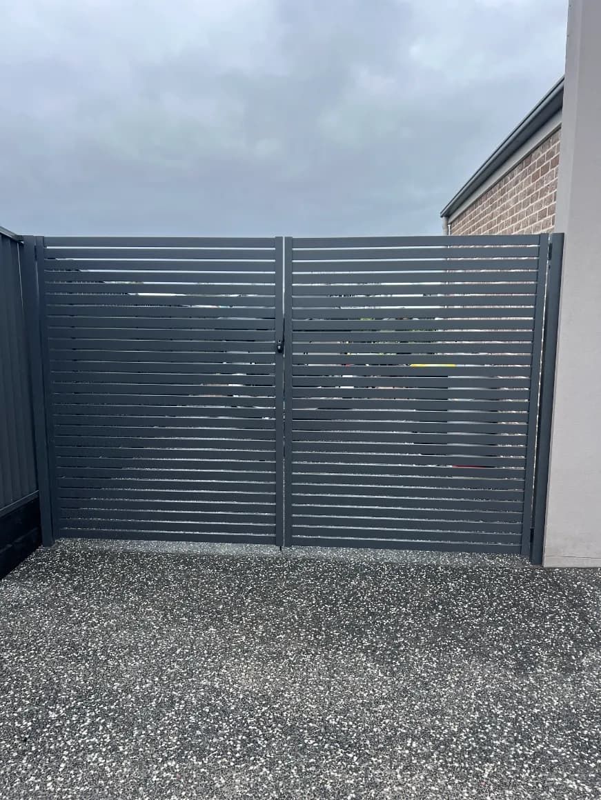 A Black Gate Is Sitting On Top Of A Gravel Driveway Next To A Brick BuildING — Mid Coast Glass Fencing In Wauchope, NSW