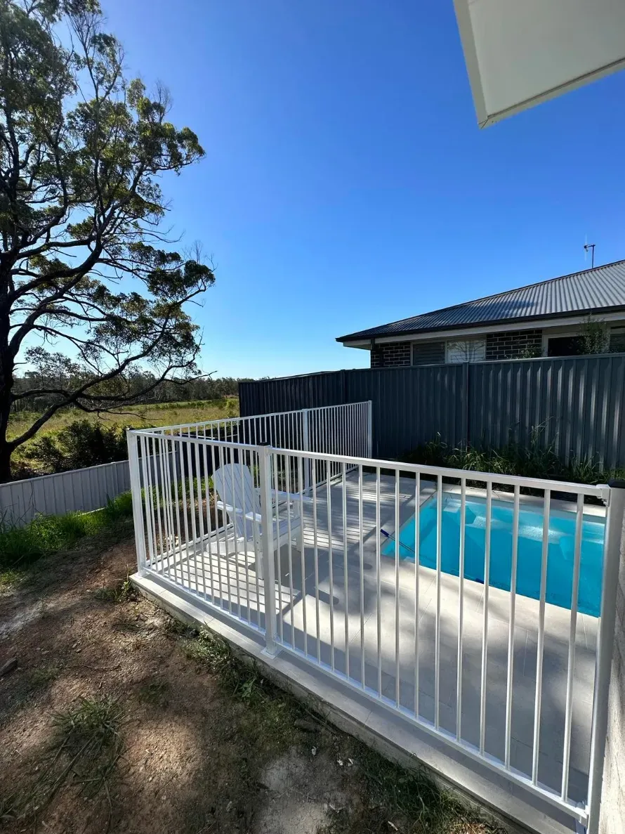 A white fence surrounds a swimming pool in front of a house — Mid Coast Glass Fencing In Wauchope, NSW