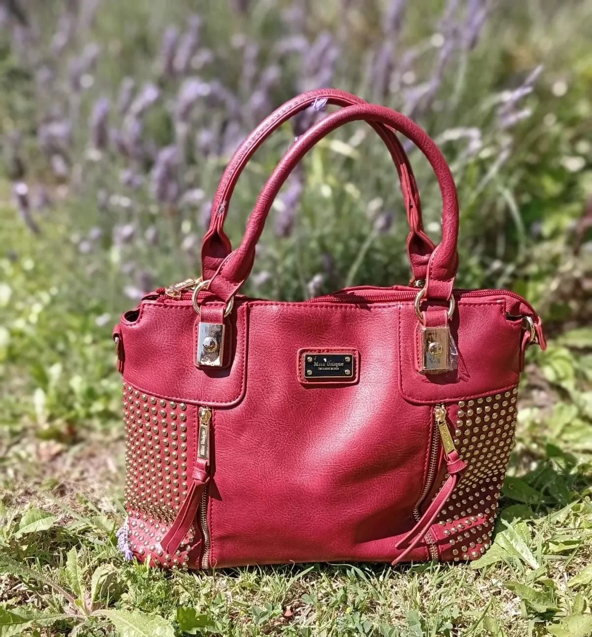 Un bolso rojo con tachuelas y detalles dorados reposa al aire libre sobre un césped, con flores de lavanda borrosas al fondo.