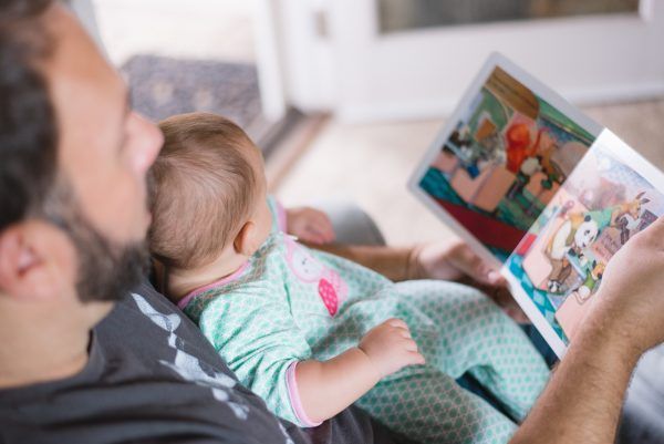 A man reading a picture book to a baby sitting on his lap.