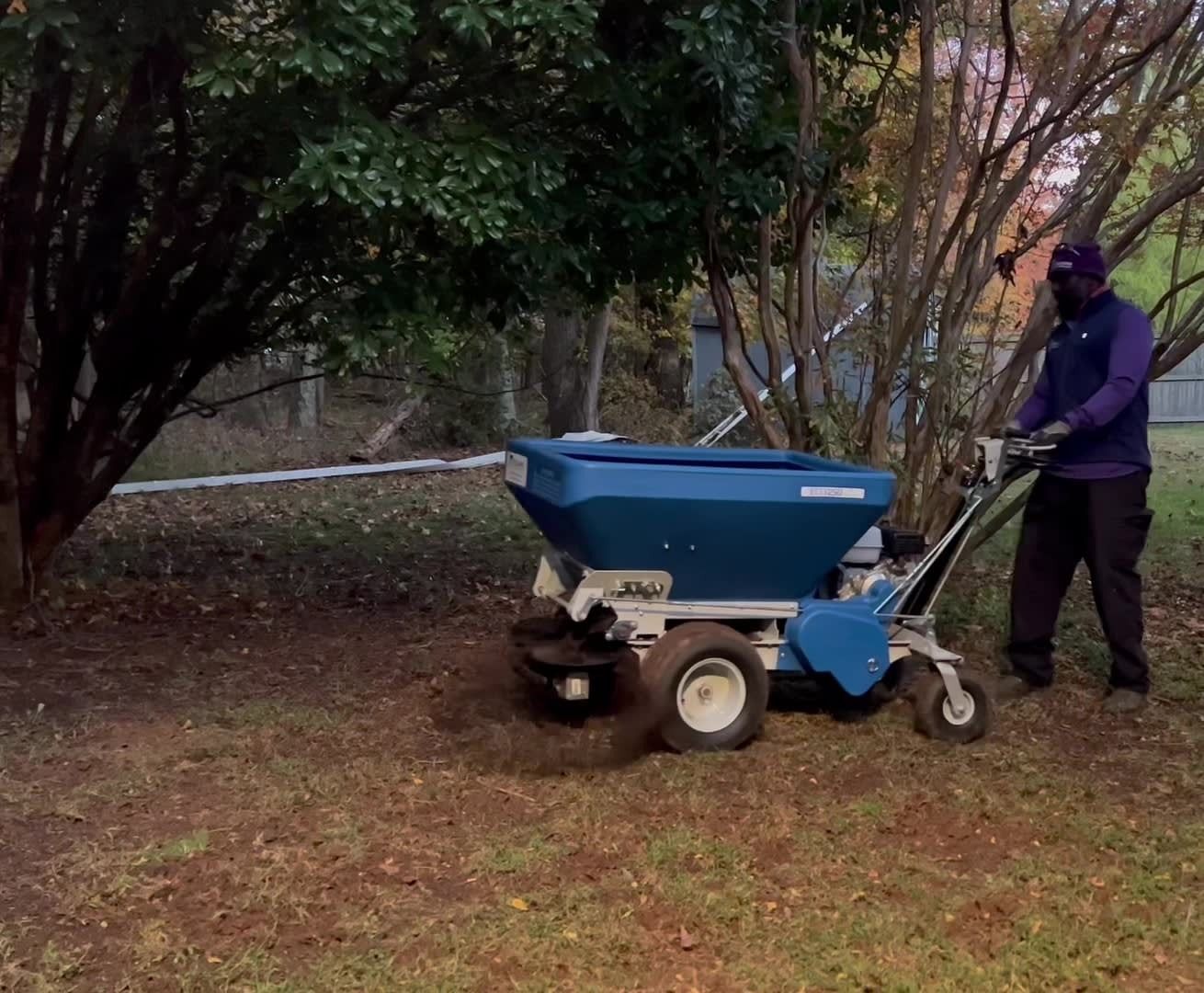 A person in a purple shirt uses a blue walk-behind lawn spreader to distribute soil or fertilizer under a tree.