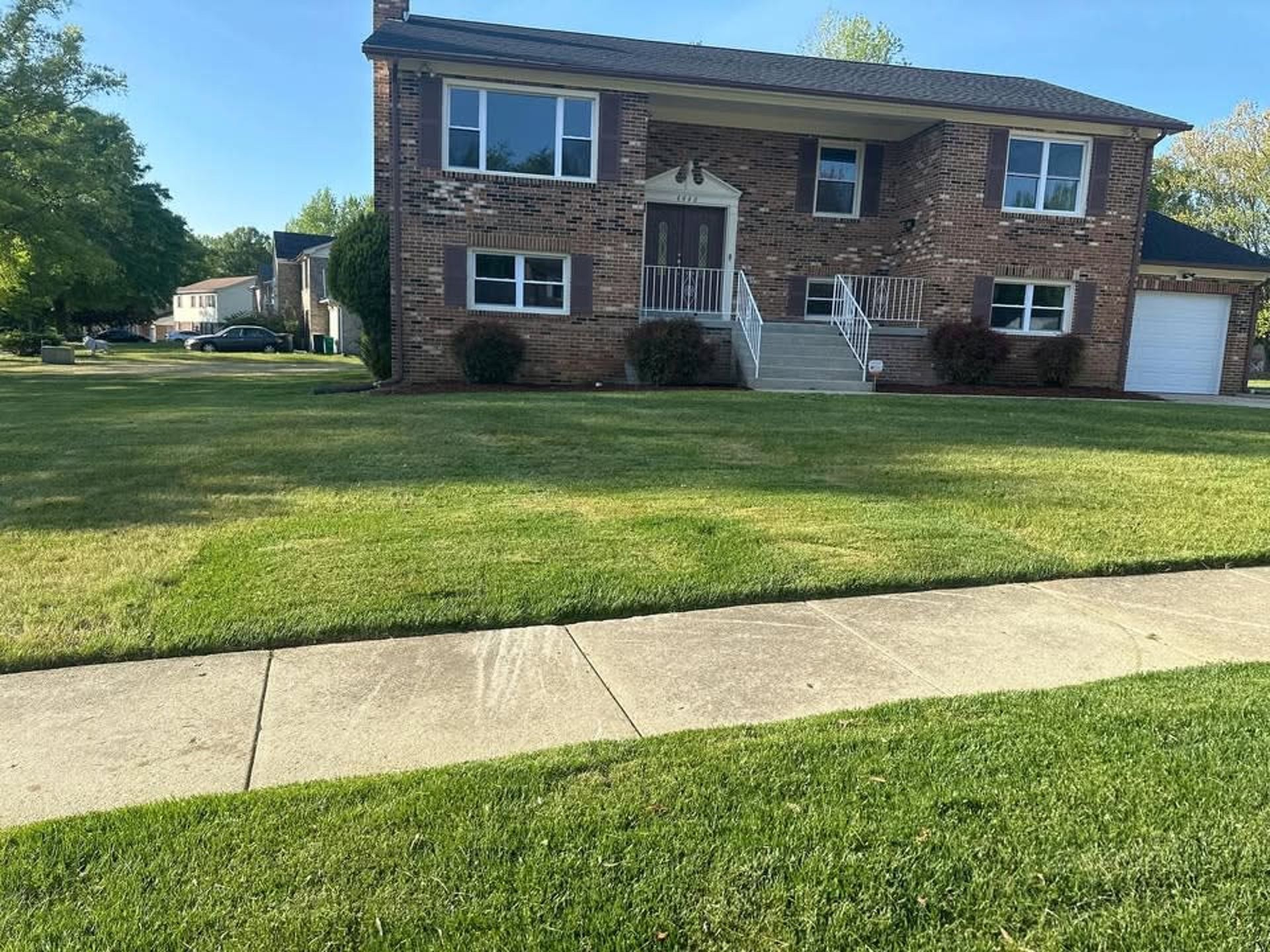 A two-story brick suburban house with a dark roof and white trim, set behind a green lawn and a concrete sidewalk.