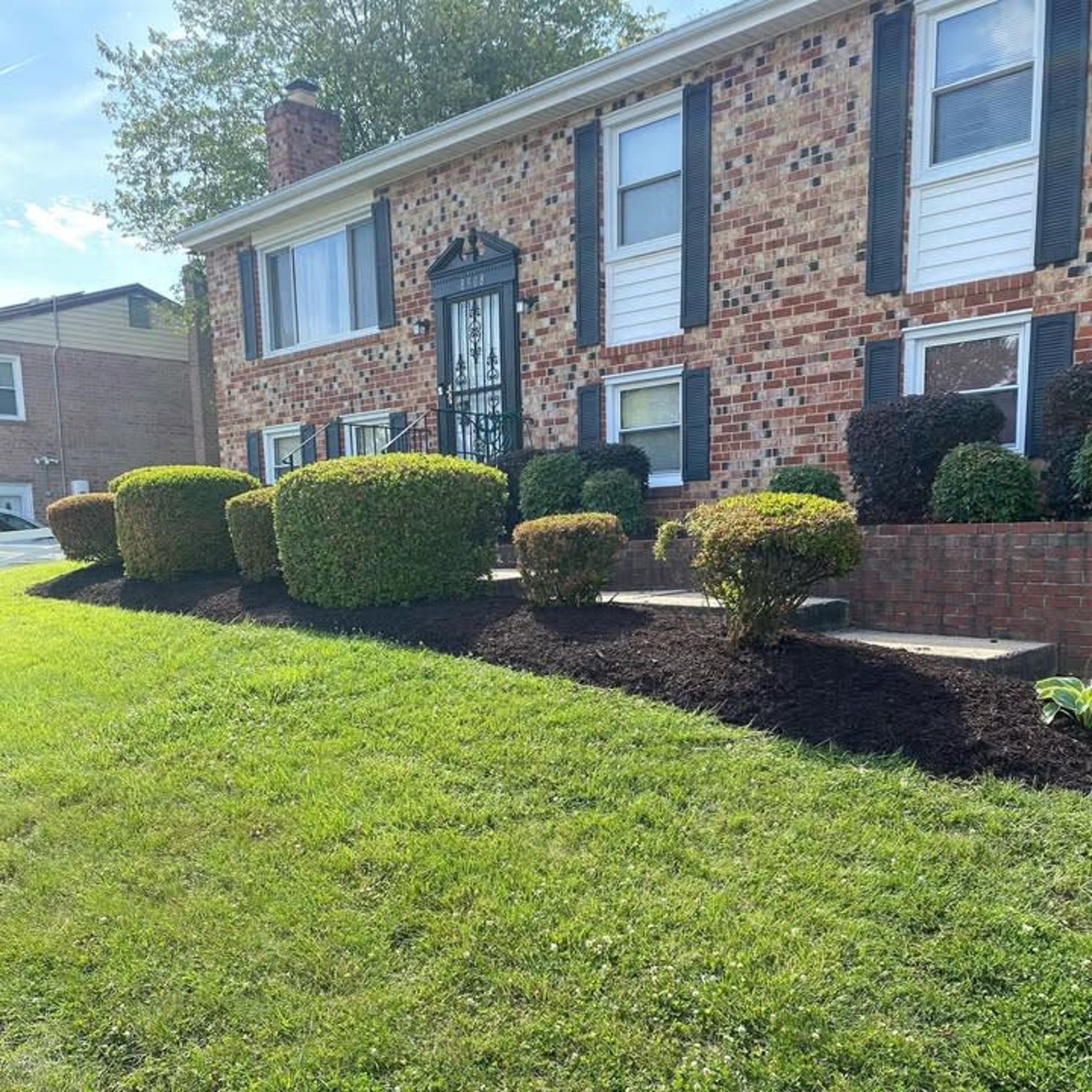 A two-story brick house with a green lawn, foundation plantings, and mulched flower beds under a clear blue sky.