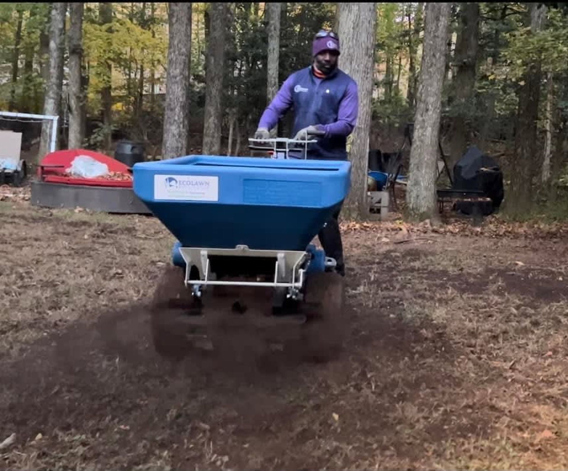 A person pushes a blue, wheeled top-dresser machine across a dirt yard, spreading soil on the ground in a wooded area.