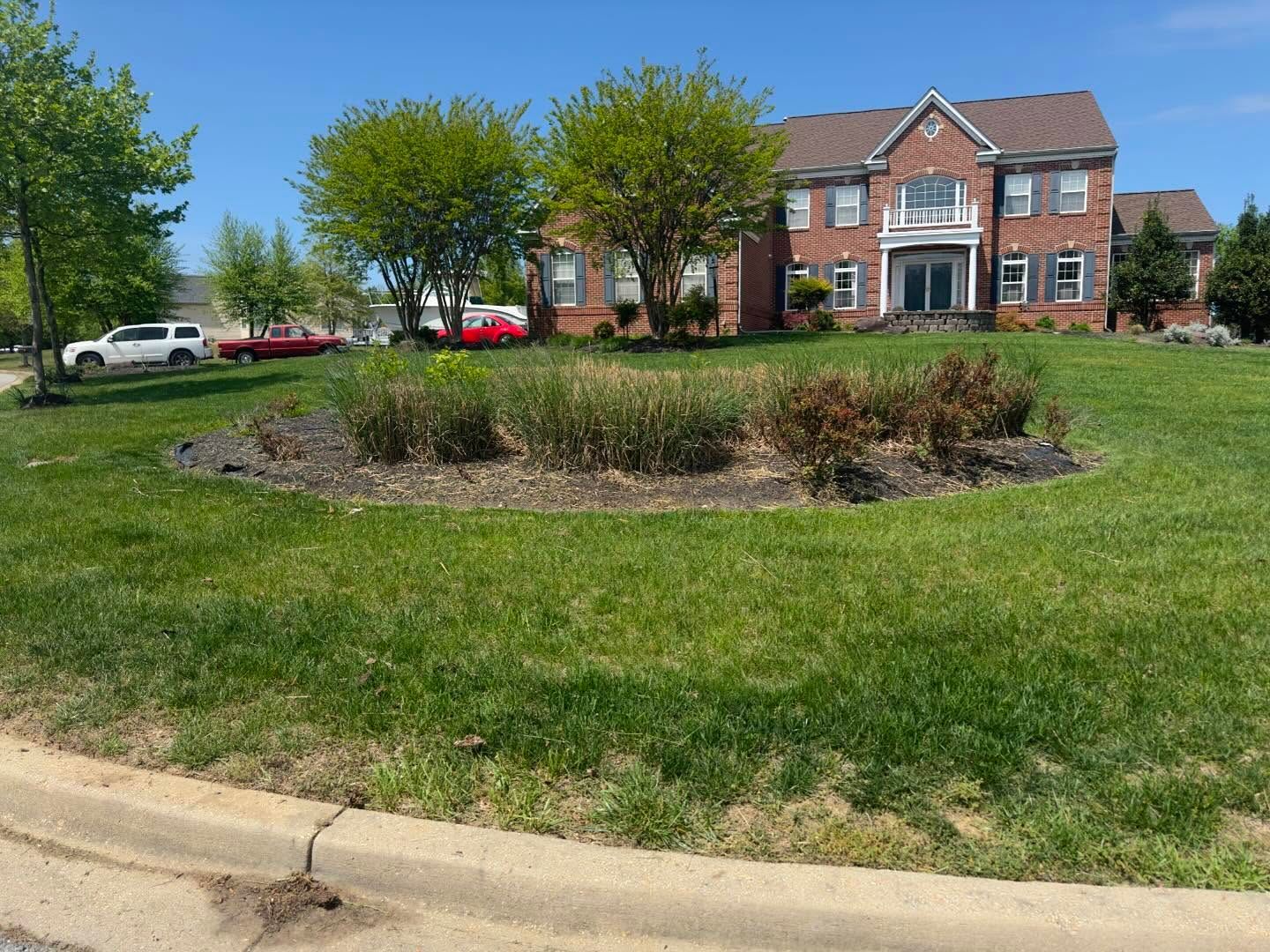 A large, two-story red brick house with a circular landscaped garden in the center of a grassy front lawn under a blue sky.