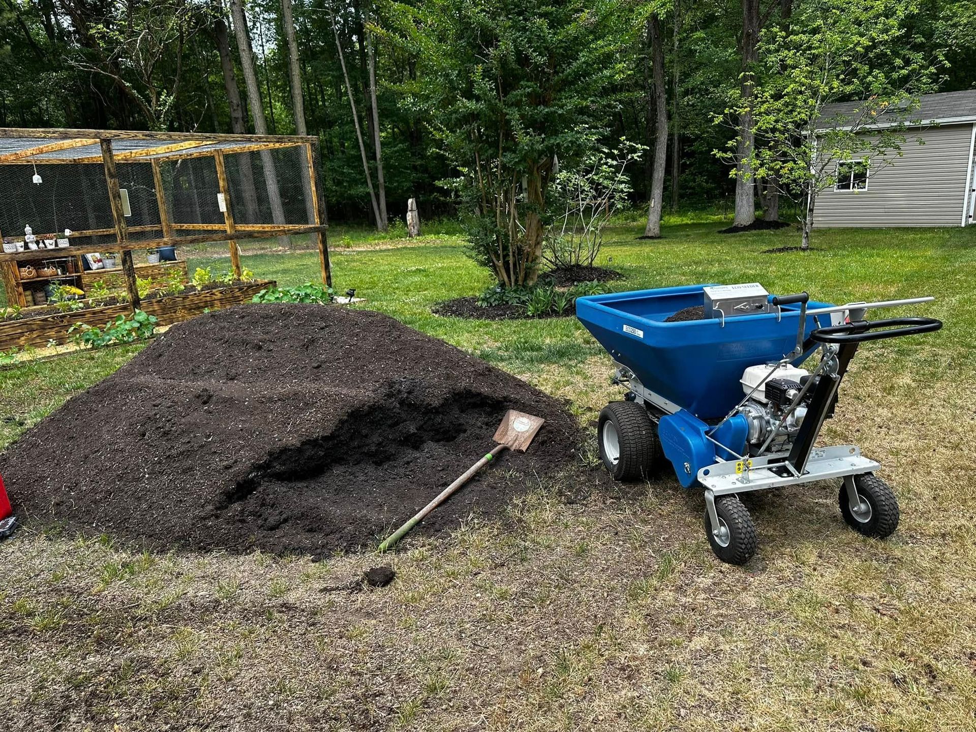 A large pile of dark mulch sits next to a blue lawn spreader in a grassy backyard near a fenced garden and shed.