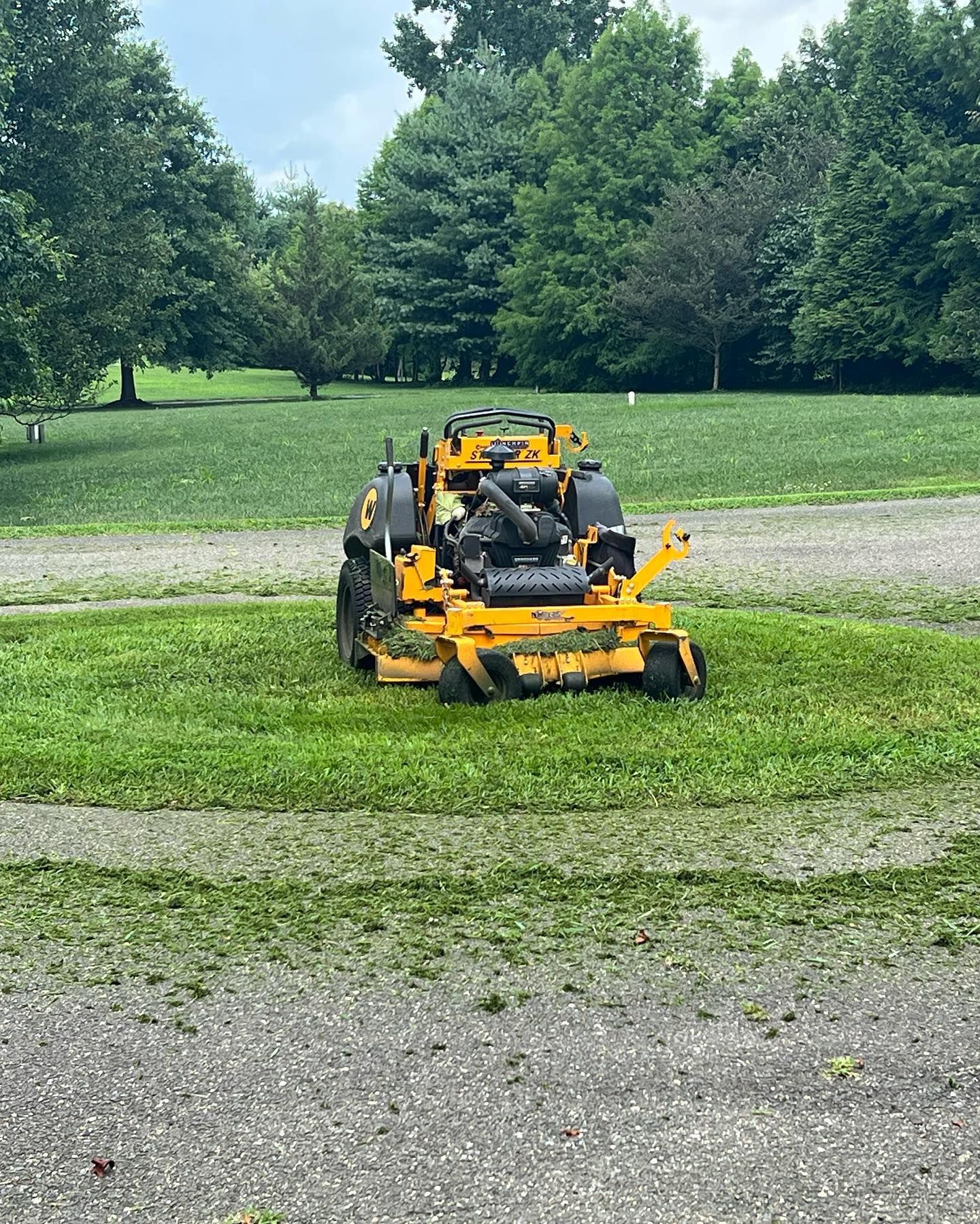 A bright yellow zero-turn lawn mower sits in the center of a circular patch of freshly cut grass in a grassy field.