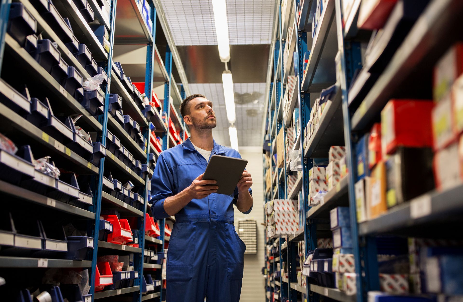 Man in blue overalls uses a tablet in a well-stocked warehouse aisle.