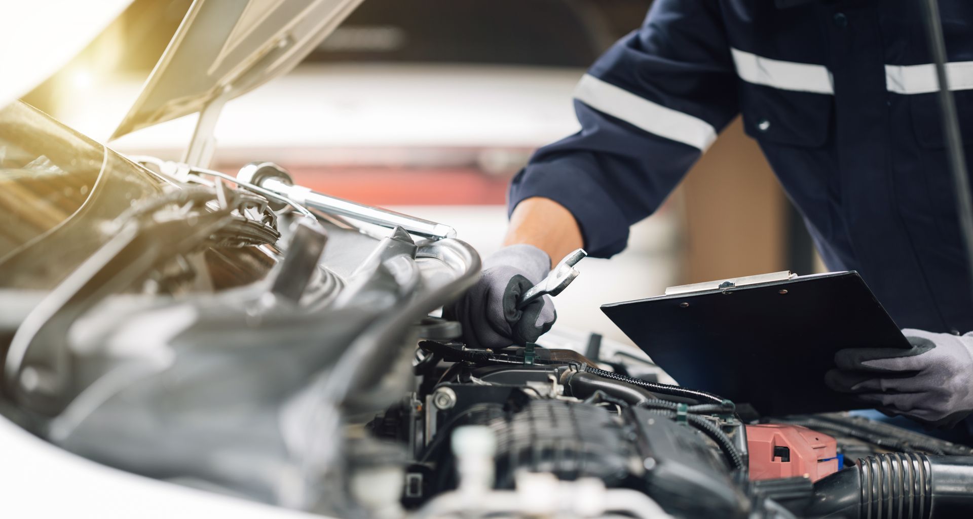 Mechanic working on car engine, holding a wrench and clipboard in a garage.