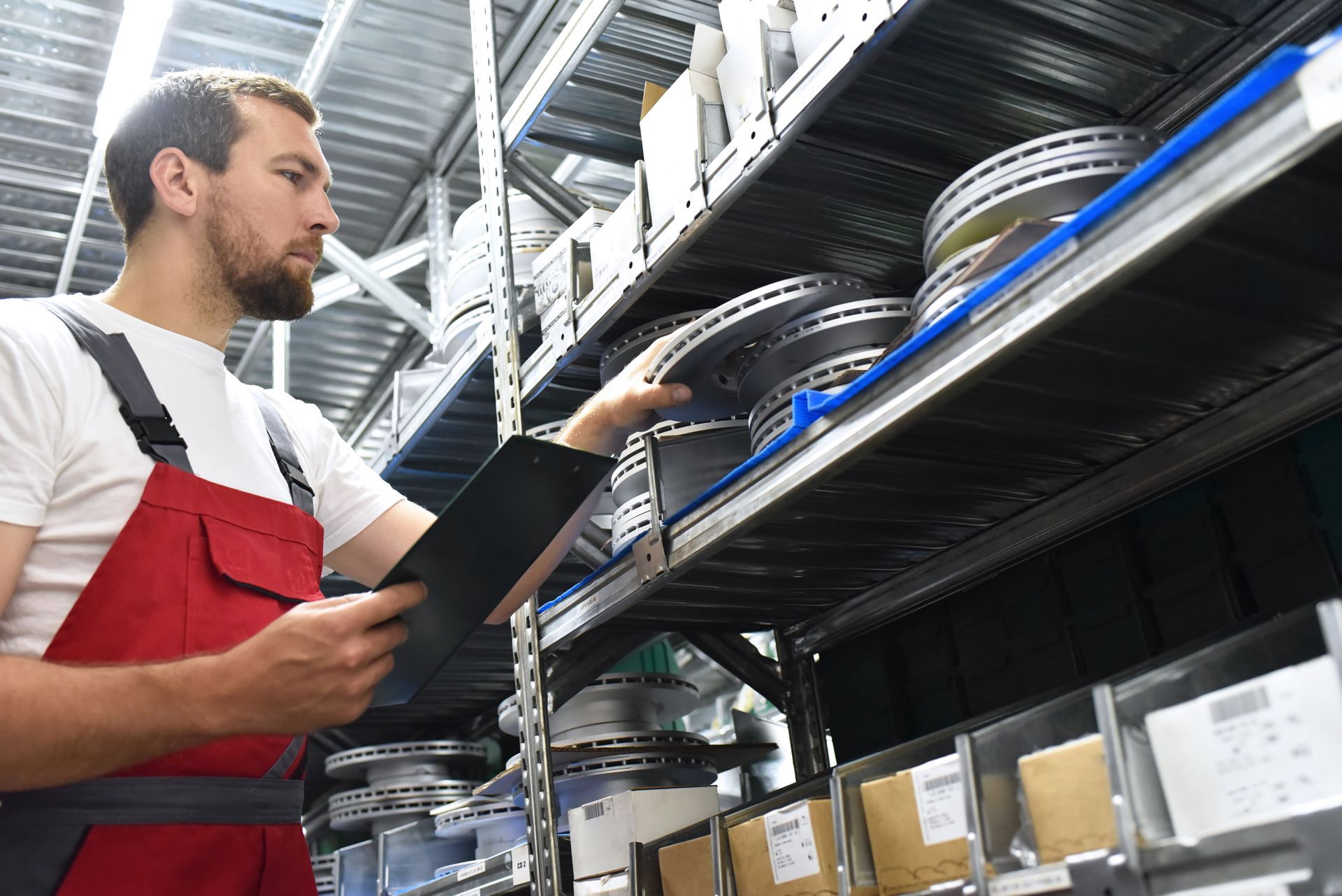 Worker in red overalls checking metal parts on storage shelves with a tablet.