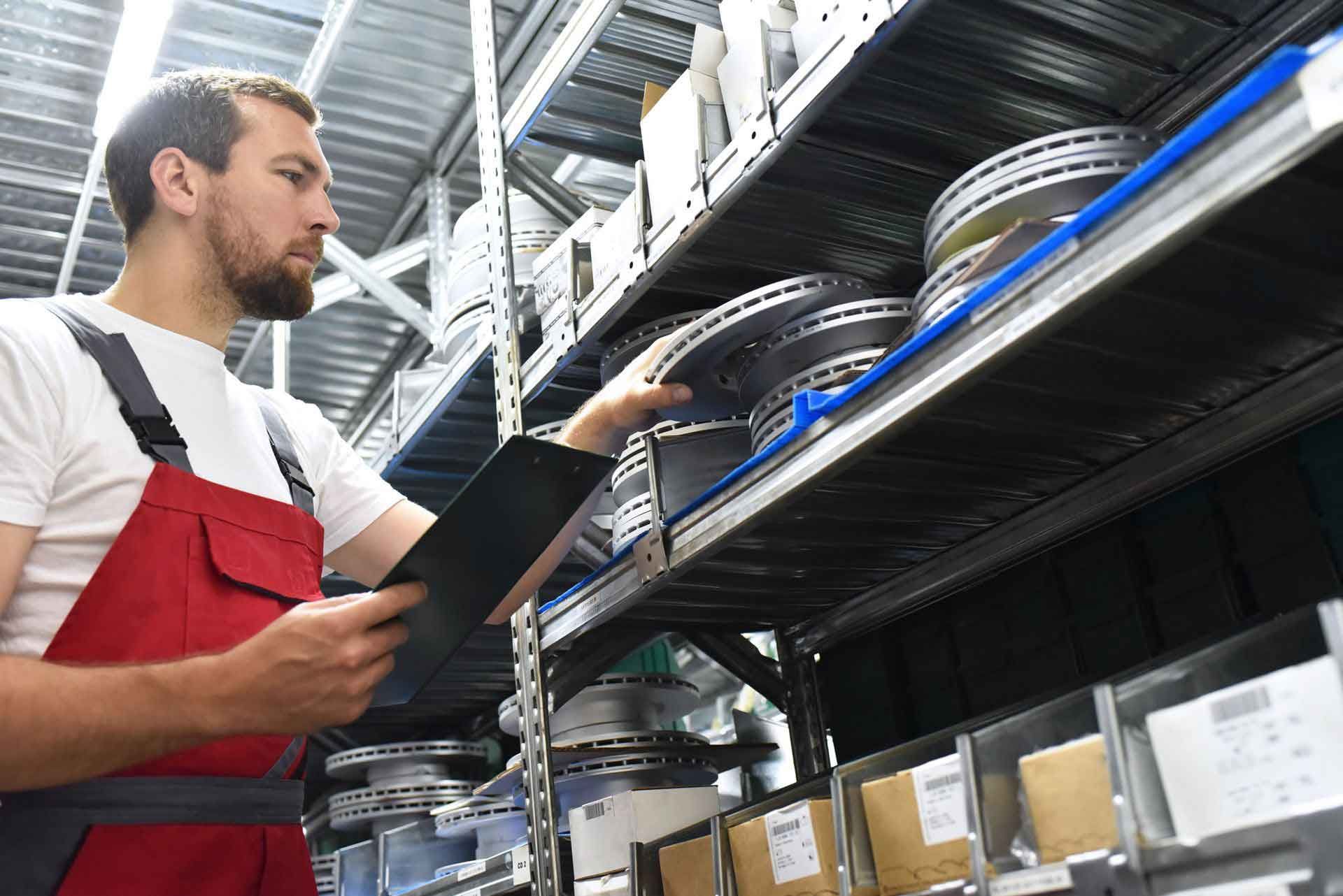 Worker in red overalls checking metal parts on storage shelves with a tablet.