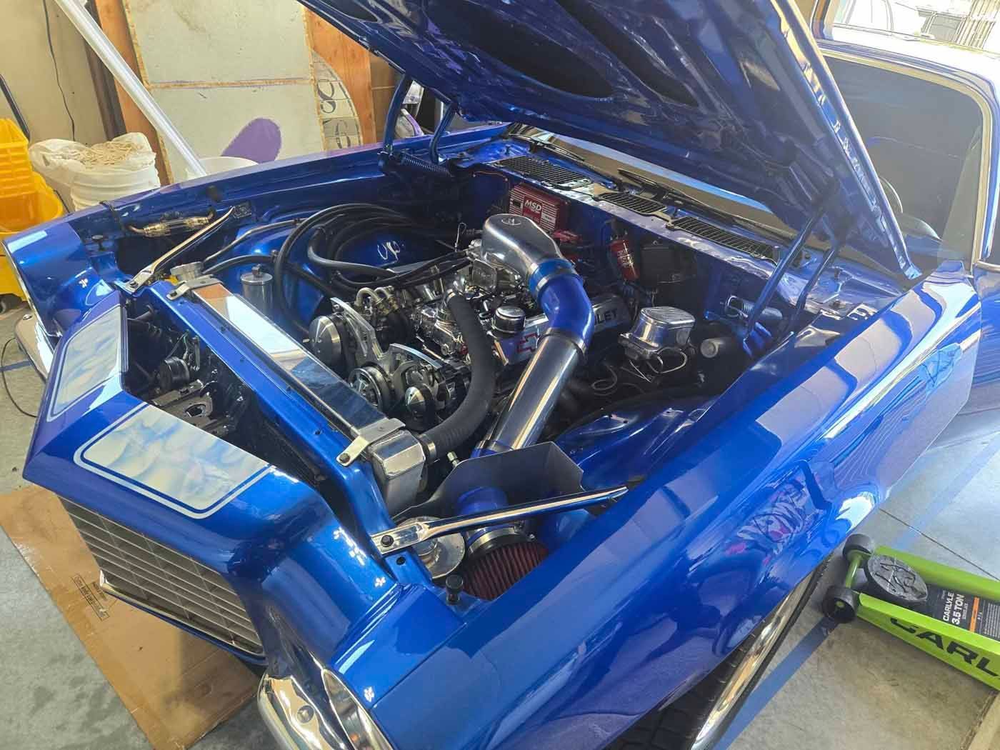 Bright blue classic car with the hood open, revealing the engine. The engine bay is customized with chrome and a blue intake.