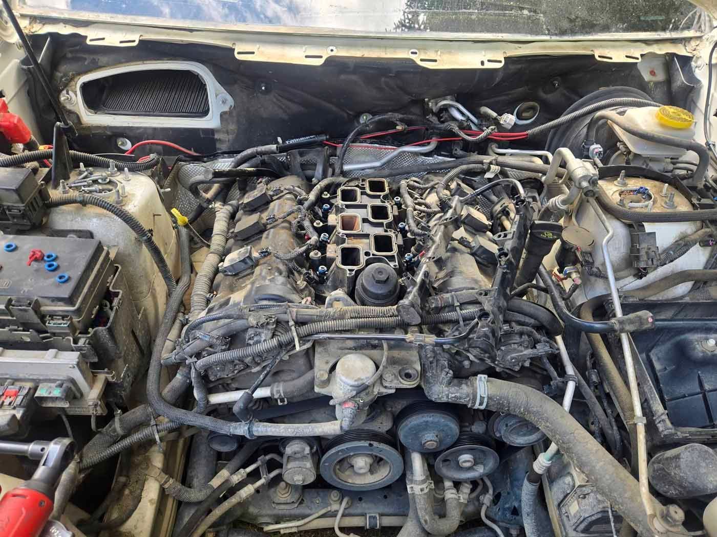Open car engine bay, with the intake manifold removed, exposing engine components. Dirty and dusty setting.